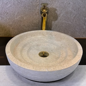 May include: A round, white marble sink with a gold drain and a gold faucet. The sink has a textured, rippled surface.