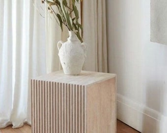 Puede incluir: Una mesa auxiliar de color beige claro con un diseño acanalado. La mesa está hecha de piedra natural y tiene un diseño simple y moderno. Un jarrón de cerámica blanco con ramas secas se encuentra encima de la mesa.