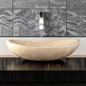 May include: A light beige oval-shaped stone vessel sink with a smooth, textured surface. The sink is sitting on a dark brown countertop.
