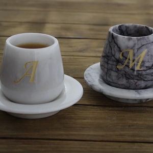 Può includere: Due tazze da caffè in marmo bianco con lettere dorate. La tazza a sinistra ha la lettera "A". La tazza a destra ha la lettera "M".