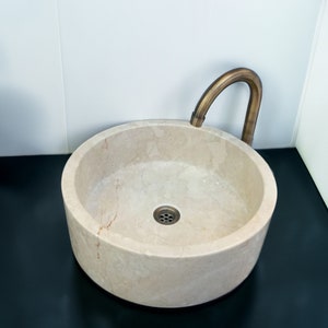 Könnte beinhalten: Ein rundes, weißes Marmorwaschbecken mit einem Messinghahn. Das Waschbecken hat einen Abfluss in der Mitte und steht auf einer schwarzen Oberfläche.