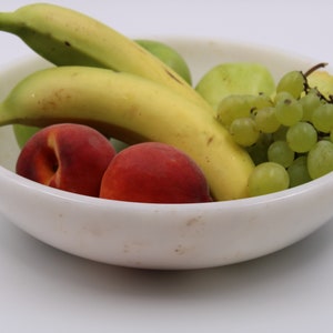 May include: A white ceramic bowl filled with fresh fruit. The bowl contains ripe bananas, green apples, red peaches, and green grapes. The fruit is arranged in a visually appealing manner, suggesting a healthy snack or centerpiece.