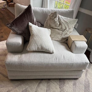 Puede incluir: Un sillón gris claro con tapicería de tela texturizada. El sillón tiene un reposabrazos de madera con una mesa auxiliar incorporada. El sillón está decorado con tres cojines: un cojín de terciopelo marrón, un cojín blanco con pliegues verticales y un cojín beige con un patrón geométrico.