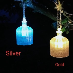 以下が含まれることがあります： 2つのボトル型の装飾用ライトが木から吊り下げられています。1つは銀色、もう1つは金色です。銀色のライトは青色に光り、金色のライトは暖かく黄色に光ります。「Silver」と「Gold」の文字が各ライトの下に書かれています。