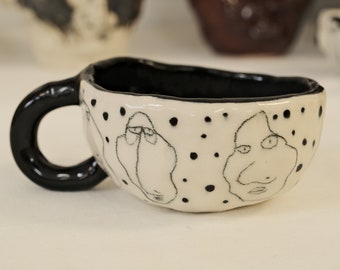 May include: A white ceramic mug with a black handle and black line drawings of faces on the outside. The mug is decorated with black dots.