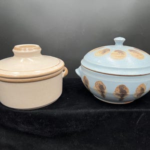 May include: Two ceramic lidded serving bowls. One is beige with brown trim and handles. The other is light blue with brown, teardrop-shaped designs. Both bowls are on a black surface.