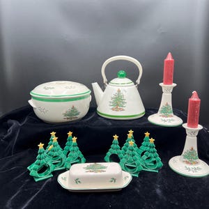 May include: A Christmas-themed porcelain tea set and tableware. The set includes a teapot, serving bowl, butter dish, candle holders with red candles, and napkin rings. Each piece is white with green trim and a Christmas tree design.