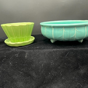 May include: Two ceramic planters. One is lime green with a fluted design and a matching saucer. The other is a turquoise oval planter with three small feet and a leaf pattern around the sides. Both are empty.