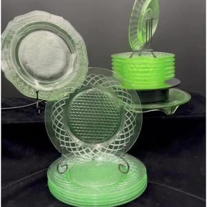 May include: A collection of vintage green glass plates. The plates have various designs, including a diamond pattern and a floral motif. Some plates are stacked, while others are displayed on stands. The glass has a slight glow.