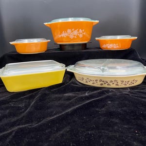 May include: A collection of vintage Pyrex casserole dishes in various sizes and shapes. The dishes feature vibrant colors like orange and yellow, with floral and leaf patterns. Each dish has a clear glass lid. The set is displayed on a black surface.