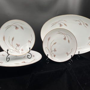 May include: A set of white porcelain dishes with a gold rim and a wheat-like design in brown and pink. The set includes a large oval platter, two round plates, and a smaller round plate. The dishes are displayed on black metal stands.