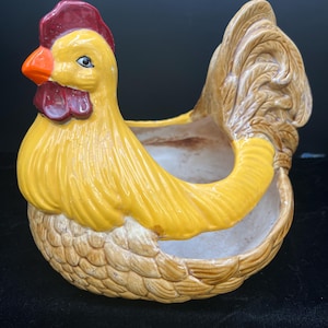 May include: A ceramic hen-shaped planter with a yellow body, a red comb and wattle, and an orange beak. The planter has a basket-like base and a hollow interior for plants or decor. The hen's tail is a light brown color.