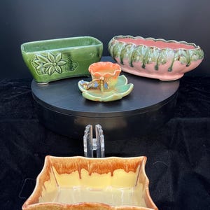 May include: A collection of ceramic planters and a small dish. The planters are in various shapes and colors, including green, pink, and yellow with brown trim. One green planter has a floral design. The small dish has a dragonfly design.