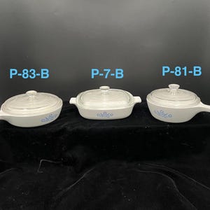 May include: Three white CorningWare dishes with clear glass lids. Each dish has a blue floral pattern and a handle or handles. The text "P-83-B", "P-7-B", and "P-81-B" is visible above each dish.