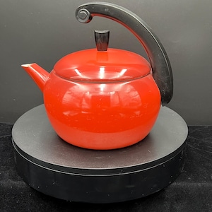 May include: A red enamel teapot with a black handle and lid knob. The teapot is sitting on a black turntable.