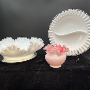 May include: A collection of white and pink decorative glassware. Includes a ruffled-edge serving bowl, a divided serving plate, and a small vase with a ruffled top. The pieces have silver or pink accents and sit on a black surface.