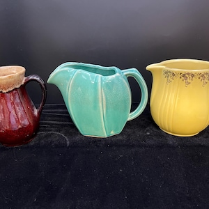 May include: Three ceramic pitchers in different colors and styles. The pitcher on the left is brown with a tan glaze. The middle pitcher is teal green and shaped like a leaf. The pitcher on the right is yellow with a gold floral design.