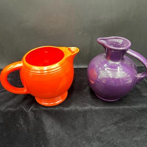 May include: Two ceramic pitchers. One is orange with a red interior, a handle, and a spout. The other is a deep purple with a rounded body, a handle, and a spout. Both are on a dark background.