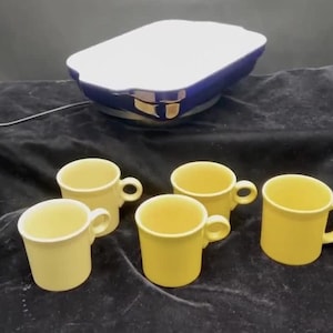 May include: A set of yellow ceramic mugs with curved handles and a blue and white casserole dish. The mugs are arranged in front of the dish on a dark velvet surface. The mugs and dish are likely for serving food.