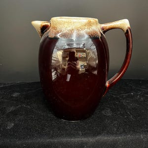 May include: A brown ceramic pitcher with a handle and spout. The pitcher has a glossy finish with a gradient of tan at the top. The handle and spout are a darker shade of brown. The pitcher is likely vintage or antique.