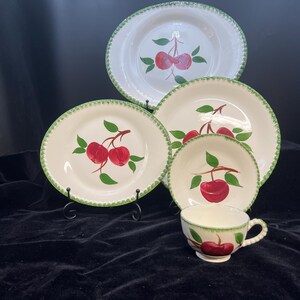 May include: A vintage white ceramic dish set with a cherry design. The set includes a large oval platter, two round plates, a small bowl, and a teacup. Each piece features a red cherry motif with green leaves and a green scalloped border.