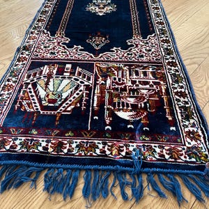 May include: A rectangular, navy blue prayer rug with intricate patterns in white, red, and gold. The rug features a central design and detailed borders, with blue fringe along the edges. The rug is placed on a wooden floor.