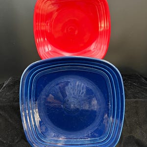 May include: Two square ceramic plates, one red and one blue, stacked on top of each other. The plates are both glazed and have a slightly raised rim.