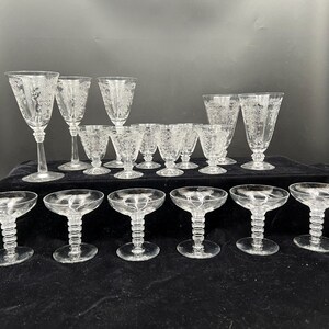 May include: A collection of clear glass stemware featuring etched floral patterns. The set includes various sizes of goblets with slender stems and wide-rimmed coupes with tiered, ribbed stems, all arranged in two rows on a dark surface.