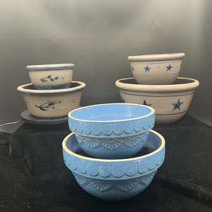 May include: A collection of ceramic bowls in various sizes and designs. Some bowls are light blue with scalloped edges, while others are beige with blue star and floral patterns. The bowls are stacked and arranged on a dark surface.