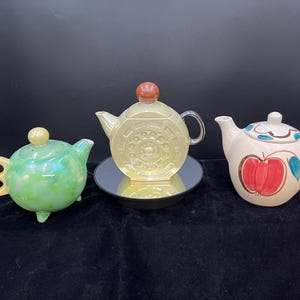 May include: Three ceramic teapots in different styles and colors. One is green with a swirl pattern, one is yellow with a circular design, and one is white with a red apple design.