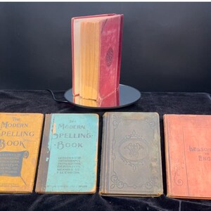 May include: Five vintage books with different colored covers. The books are titled "The Modern Spelling Book", "The Modern Spelling Book", "Lessons in English", and two books with no visible titles. The books are arranged in a row on a black surface.