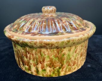 May include: A round, lidded ceramic dish with a speckled pattern in shades of brown, green, and tan. The lid has a small knob and a ridged design. The dish is likely for serving or storing food.
