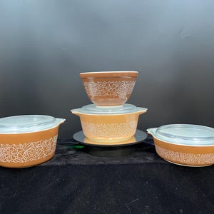 May include: A collection of vintage Pyrex bowls and casserole dishes in shades of brown and orange. Each piece features a white floral pattern and clear glass lids. The bowls are stacked, showcasing their different sizes and shapes.