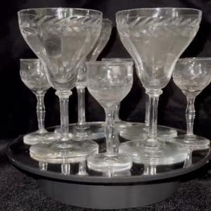 May include: A set of clear, etched glass goblets and smaller glasses arranged on a mirrored surface. The glasses have a delicate, floral-like pattern around the rims and are displayed against a dark background, highlighting their transparency and design.