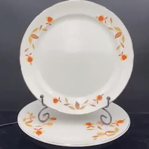 May include: White ceramic plates with a floral design in orange, yellow, and brown. The plates are displayed on a decorative metal stand. The floral pattern is arranged around the edges of the plates.