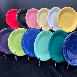 May include: A set of 12 colorful ceramic dinner plates displayed on a black metal stand. The plates are in various shades of blue, green, yellow, pink, purple, and black.