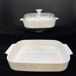 May include: Two white rectangular casserole dishes with a floral iris design. One dish has a clear glass lid with a clear handle. The dishes are decorated with purple and green iris flowers. The dishes are on a black surface.