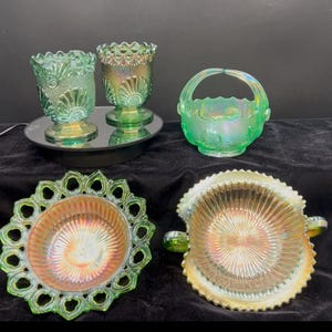 May include: An assortment of iridescent green glass decorative items. The collection includes two stemmed goblets, a small basket with a handle, and two decorative bowls. The glass has a textured, patterned design. The items are displayed on a black surface.