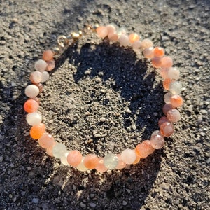 May include: A bracelet made of small, round, orange, white, and clear sunstone beads. The bracelet is on a gray, textured surface.