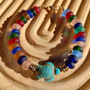 May include: A colorful beaded bracelet featuring a turquoise turtle charm. The bracelet is made of round, frosted glass beads in various colors, including red, blue, green, and pink. The bracelet is secured with a gold-colored clasp.