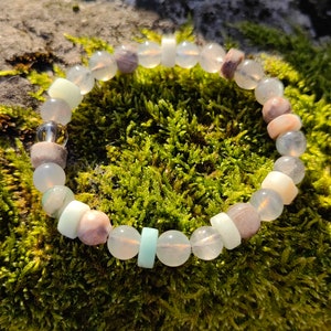 May include: A bracelet made of light pink, white, and light green beads. The bracelet is on a bed of green moss.