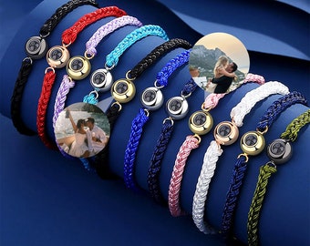 May include: A collection of personalized bracelets with woven bands in various colors, including black, red, and blue. Each bracelet features a round charm with a photo and a metallic accent, displayed on a blue surface.