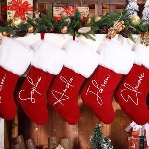 May include: Red Christmas stockings with white faux fur trim, each personalized with a name in elegant script. The stockings are hanging above a fireplace decorated with festive greenery and ornaments.