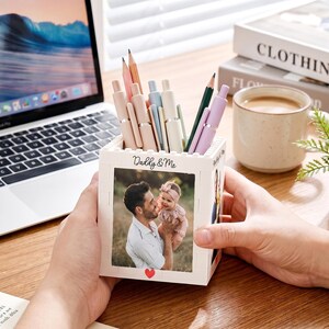 Peut inclure: Un porte-stylo carré blanc à la texture en blocs, présentant une photographie imprimée d'un homme tenant un bébé sur le panneau avant. Le texte Daddy & Me est imprimé au-dessus de la photo et un petit cœur rouge est imprimé en bas au centre.