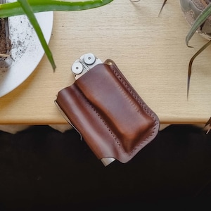 Może przedstawiać: Brązowa skórzana pochwa na multi-narzędzie z częściowo widocznym multi-narzędziem ze stali nierdzewnej. Pochwa jest szyta i ma prosty design. Zdjęcie zrobione z góry.