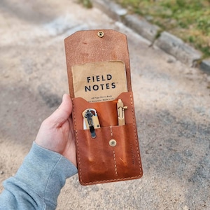 May include: A brown leather notebook cover containing a brass pen, a small knife, and a Field Notes memo book. The cover is open, showing the contents. The cover is held in a person's hand.