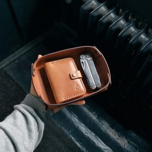 May include: A tan leather tray containing a tan leather wallet with a button clasp and a silver multi-tool. The tray and wallet are light brown. The multi-tool is silver and has various tools. The items are held in a person's hand.