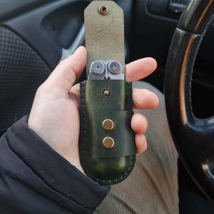 May include: A green leather multi-tool case with a snap closure and two brass-colored buttons. The case holds a silver multi-tool with various implements. The case is open, revealing the tool.