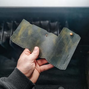 May include: A handcrafted, olive green leather wallet with a unique shape. The wallet features a button closure and visible stitching along the edges. The text "WANDERLUST LEATHER CO." is stamped on the side. The leather has a distressed, textured appearance.