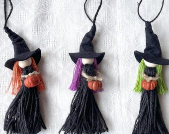 May include: Three handmade Halloween witch ornaments. Each witch has a black felt hat, a wooden bead face, and a black yarn dress. One has orange hair, one purple, and one green, each holding a small pumpkin.